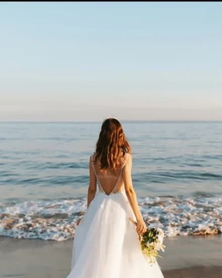 A bride in a white dress walking towards the sea at golden hour, holding her bouquet behind her.
